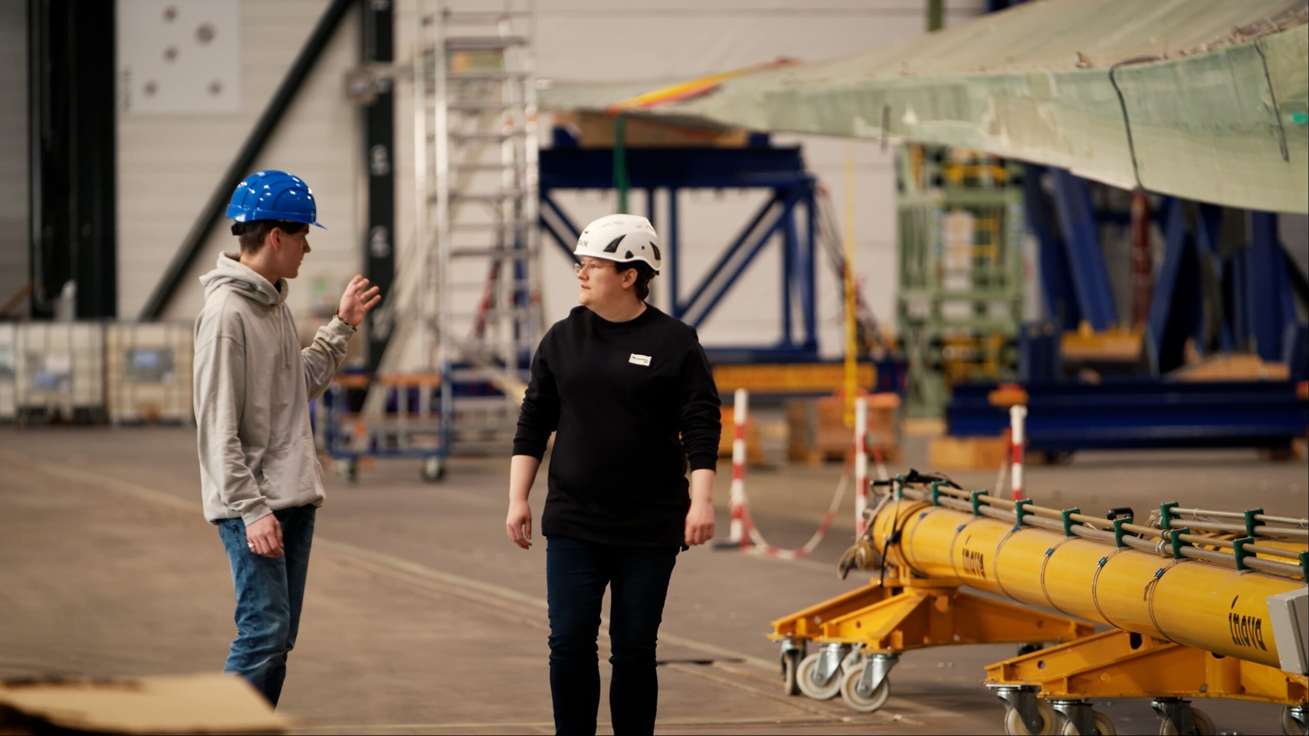 Rabanna Urspruch, Rotorblattprüferin am Fraunhofer IWES, im Austausch mit Schüler Jendrik Döhring. Beide stehen in einer Halle. Im Hintergrund sind Leitern, Rotorblätter und Maschinen.